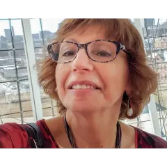 I don't know who this person is, but it shows a woman with glasses smiling indoors near a window.